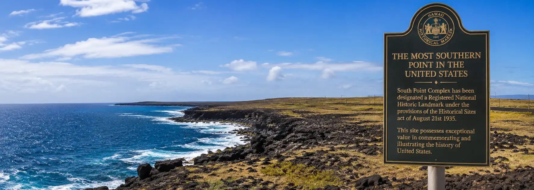 The Southern Most Point of the United States of America Historic marker at South Point on the Big Island of Hawaii overlooks rugged volcanic cliffs and the Pacific Ocean, marking the southernmost point in the United States beneath a bright tropical sky.
