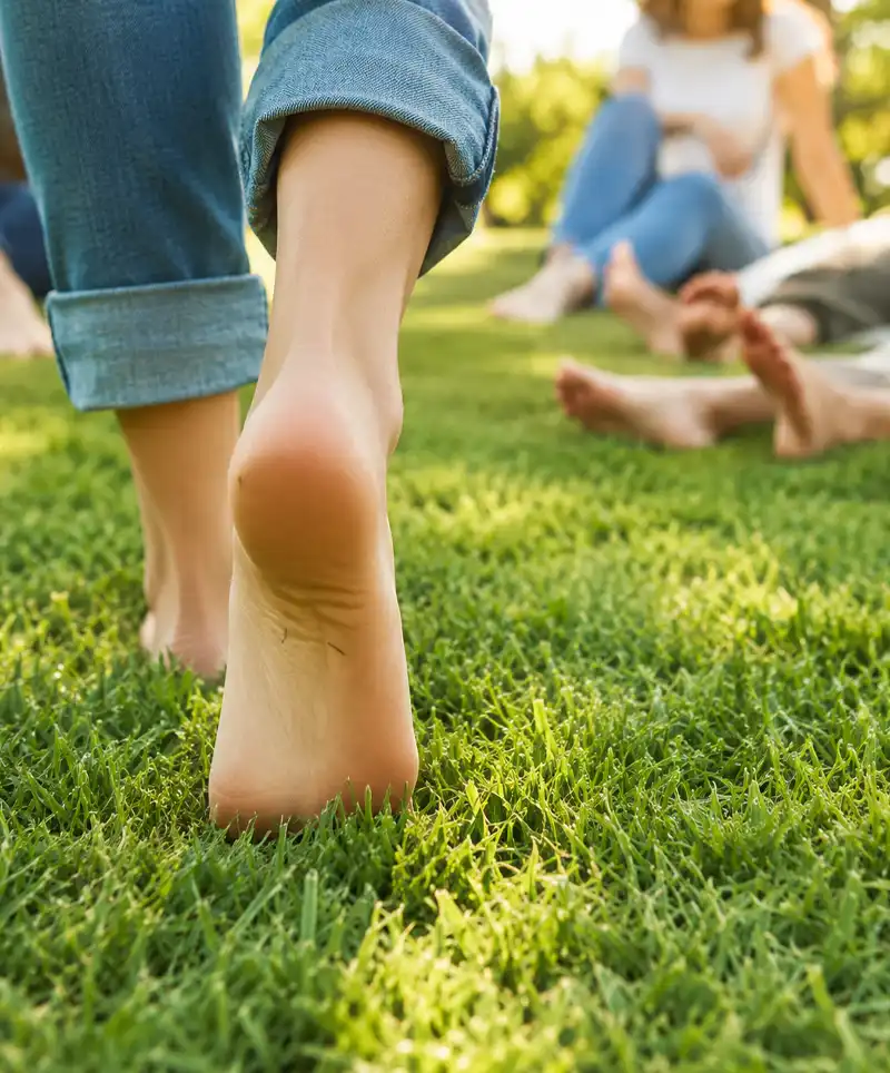 Close-up of bare feet walking through soft green grass while others relax barefoot in the background, highlighting the simple joy of walking barefoot in nature on a warm sunny day.
