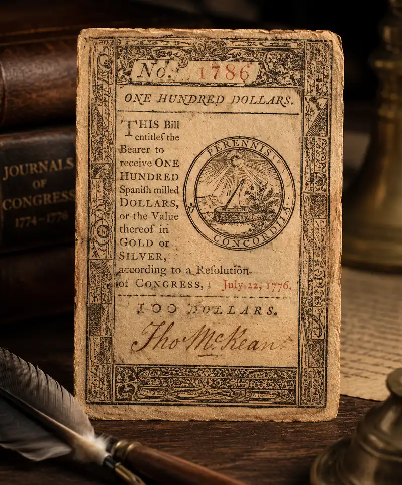 Antique-style American banknote displayed on a wooden desk beside a quill pen, ink well, and old books, evoking the early history of U.S. paper currency and the origins of National Dollar Day.