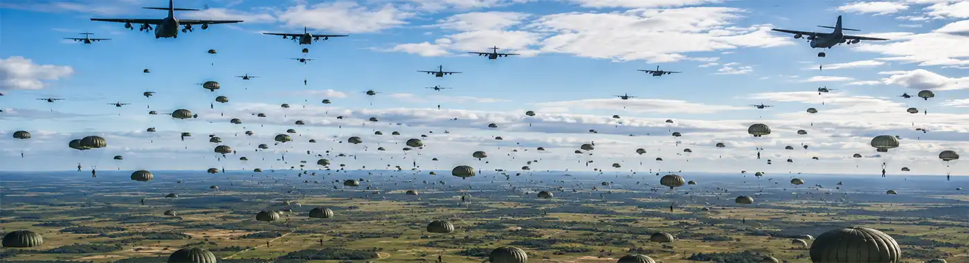Hundreds of U.S. Army paratroopers descend beneath round parachutes during a large-scale daytime training exercise as multiple military transport aircraft fly over an expansive drop zone on a military base.
