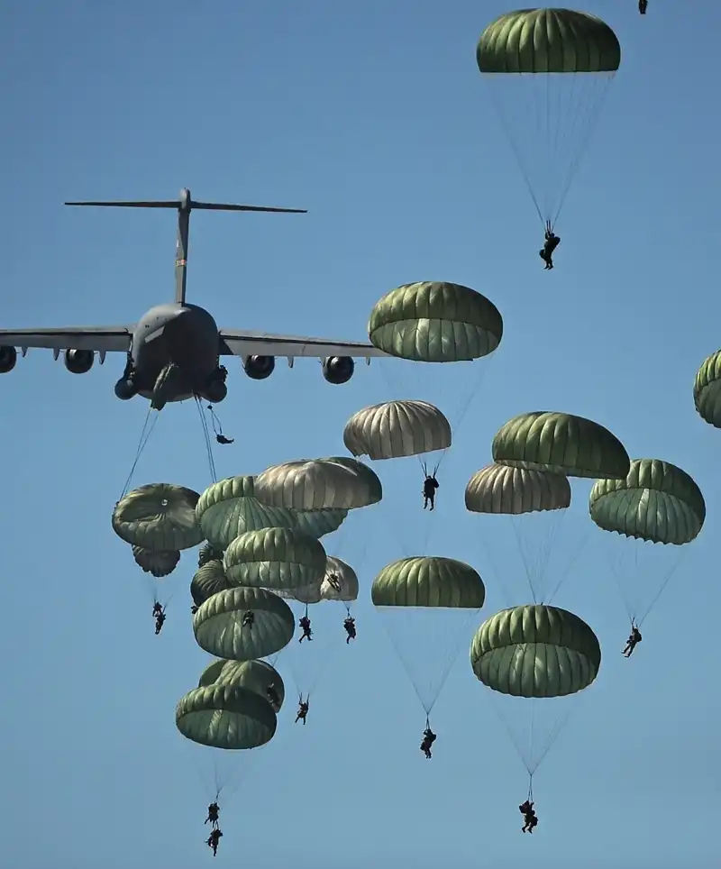 A string of U.S. Army paratroopers exits a military transport aircraft during a dawn airborne operation, with dozens of parachutes descending toward the drop zone below under the rising sun.