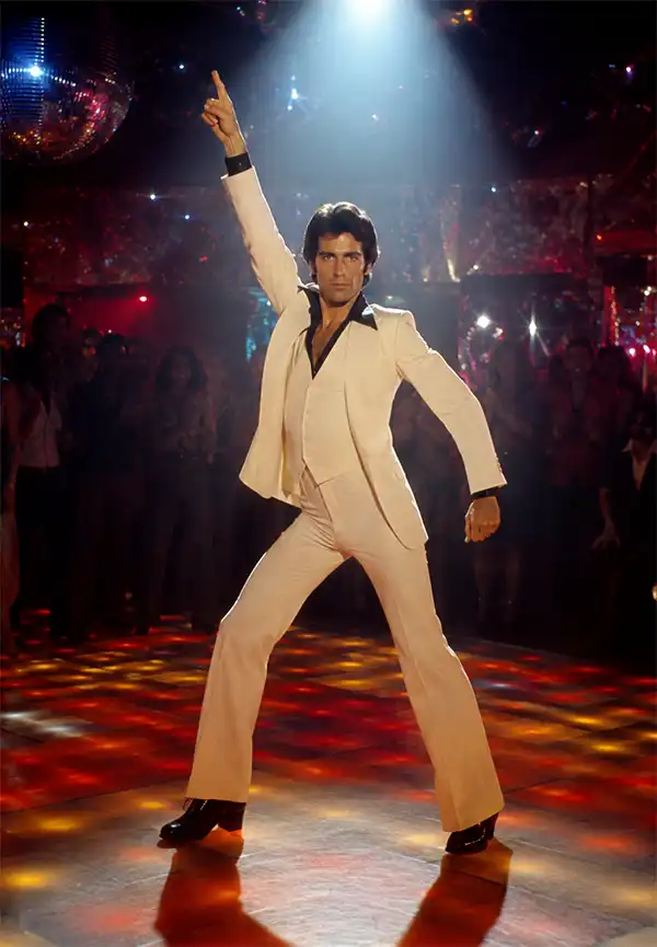 Oh, come on. You know we had to do it. A male disco dancer in classic 1970s attire strikes a dramatic pose beneath a spotlight on a glowing dance floor, surrounded by colorful lights, mirror balls, and an enthusiastic nightclub crowd.