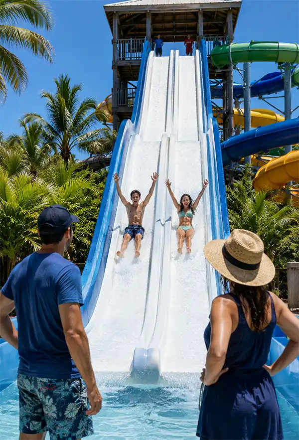 Two teenagers race down a tall water slide with their arms raised while their parents watch from the pool below.