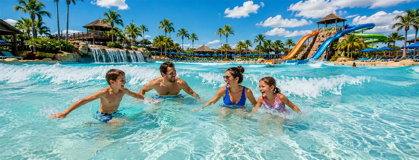 Family laughing and playing together in a sunny waterpark wave pool, surrounded by palm trees, waterfalls, and colorful water slides.