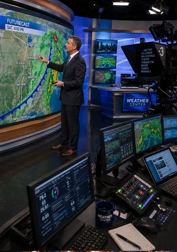 A television weather forecaster presents a live forecast in a modern broadcast studio, viewed from behind the scenes with cameras, monitors, control consoles, and advanced computer systems used to analyze and display weather data.