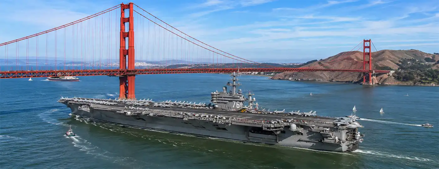 A U.S. Navy aircraft carrier sails beneath the iconic Golden Gate Bridge on a clear, sunny day, escorted across calm waters with its flight deck lined with aircraft against the beautiful San Francisco skyline.