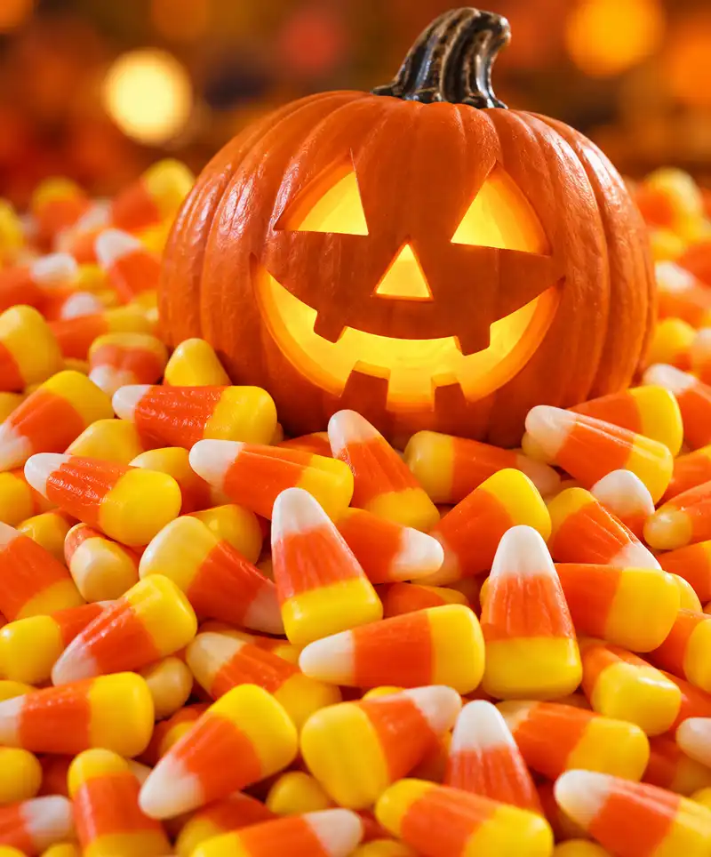 A glowing, smiling jack-o'-lantern sits atop a large pile of yellow, orange, and white candy corn, surrounded by warm autumn colors in a festive Halloween setting.